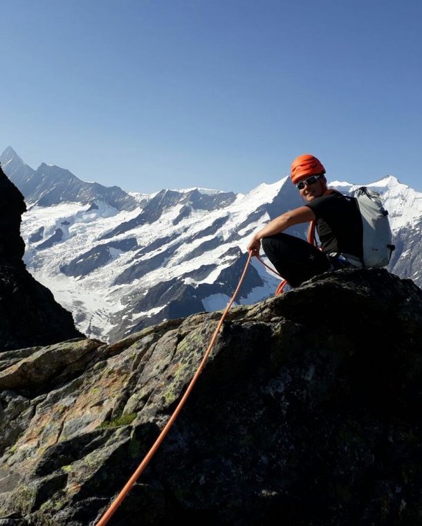 Climber in helmet and harness on rocky peak, snowy mountains in background, holding a rope.