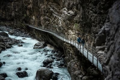 Two people with backpacks and helmets are hiking along a narrow path next to a rock wall above a raging river.