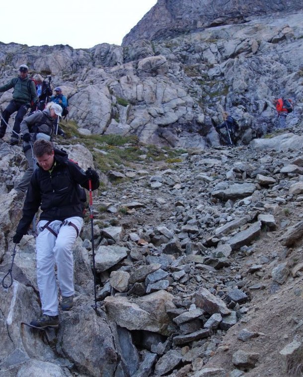 People with helmets and climbing harnesses are descending a rocky mountain path using poles.