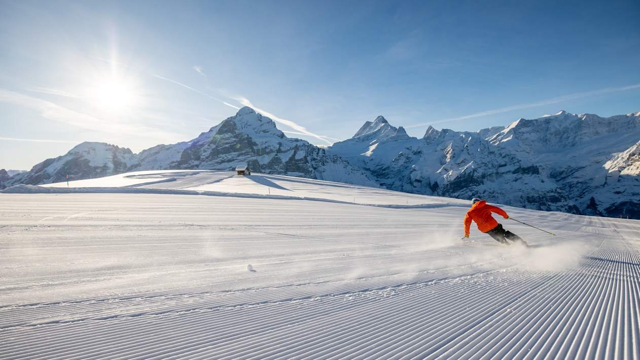 Person skiing down a groomed slope, surrounded by snow-covered mountains.