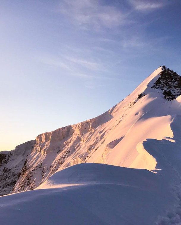 Snow-covered mountain peak at sunrise with a clear sky, showing footprints along the ridge.