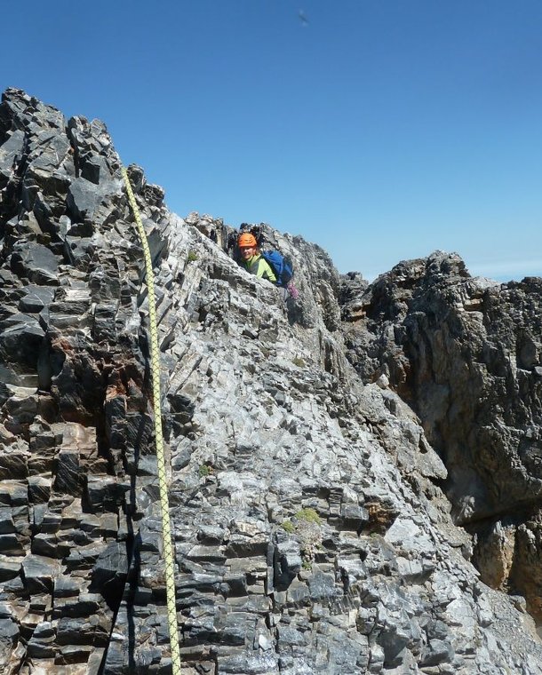 Climber with helmet and harness ascends rocky mountain ridge, secured by rope, under clear blue sky.