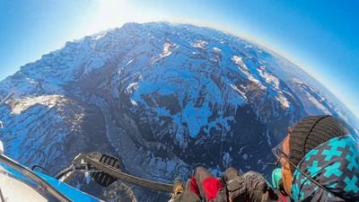 People with helmets and winter clothing are sitting in a helicopter over snow-covered mountains.