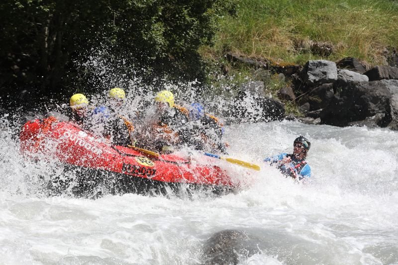 People in yellow helmets are rafting in a red boat through whitewater, surrounded by rocks and trees.