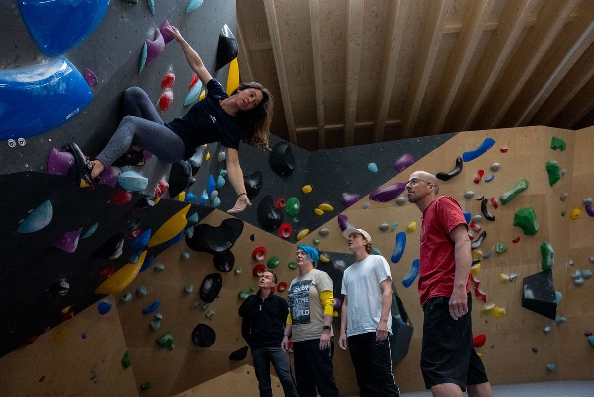 Person climbing an indoor bouldering wall with colorful holds, observed by four others in casual attire.