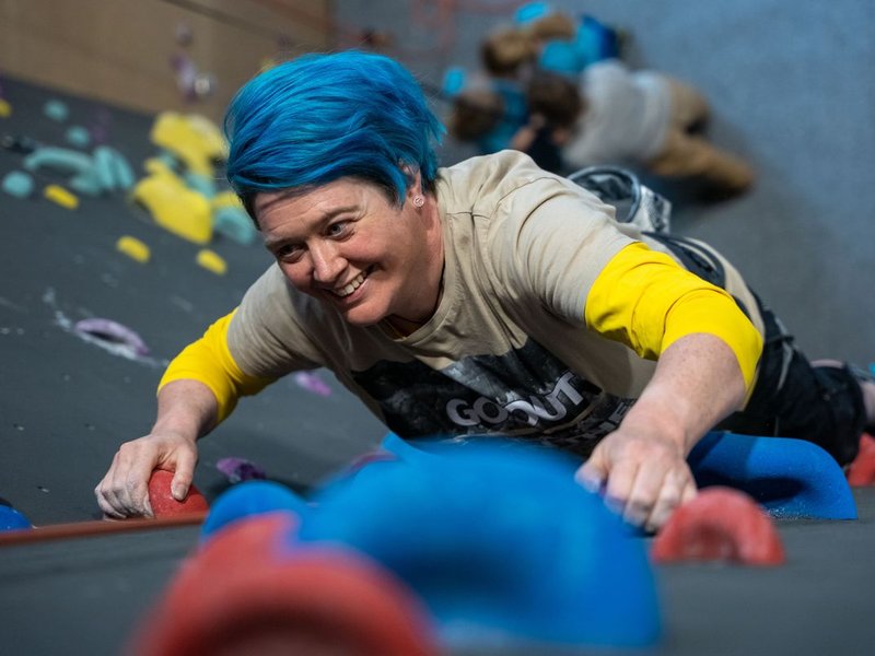 Person with blue hair climbing an indoor wall, wearing a harness and casual clothing.