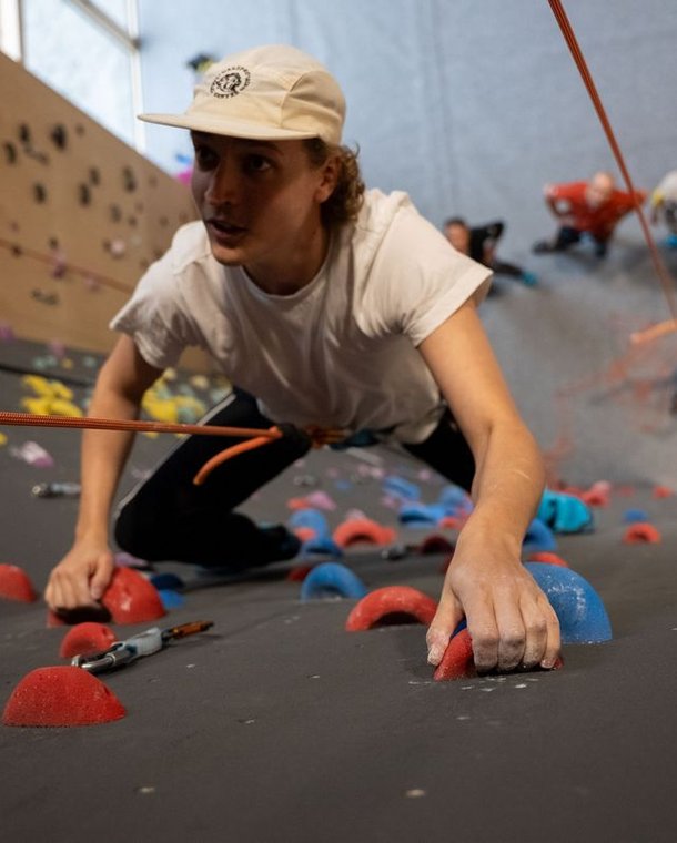Person climbing indoor wall with colorful holds, wearing a white shirt, cap, and harness with rope.