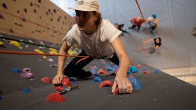 Person climbing indoor wall with colorful holds, wearing a white shirt, cap, and harness with rope.