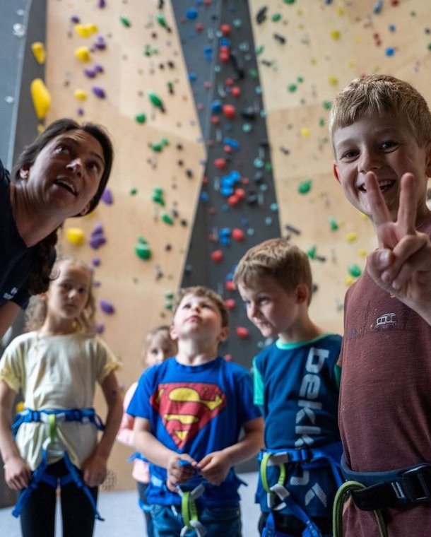 Children in harnesses listen to an instructor at an indoor climbing wall.