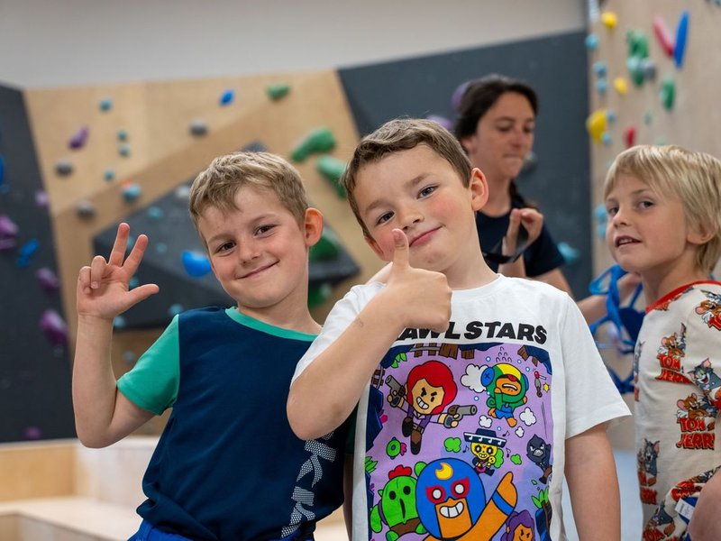 Children in a climbing gym with colorful holds on the wall; an adult is in the background.