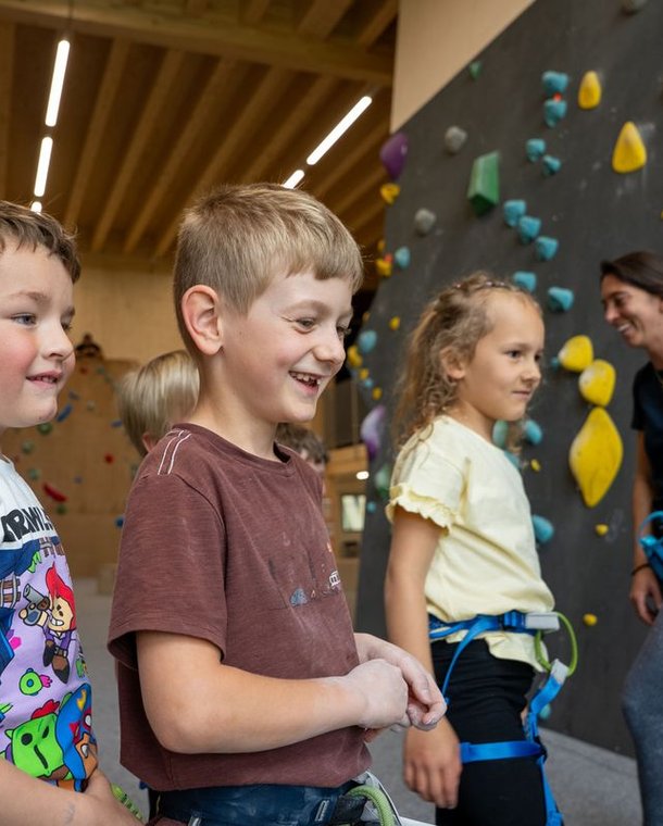 Children with harnesses in an indoor climbing gym, colorful holds on walls, adult guide present.