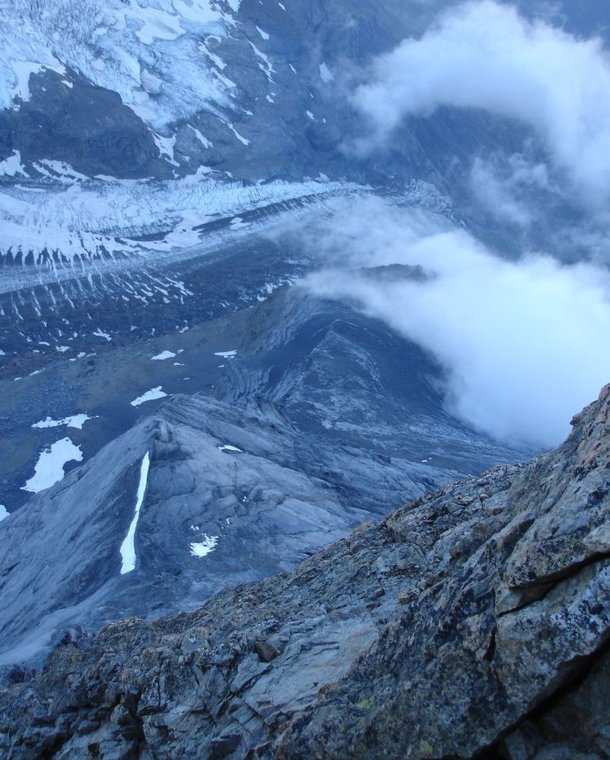 Rocky mountain peak with snow patches and clouds, overlooking a glacier and rugged terrain below.