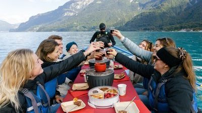 Group on a boat in life jackets toasting drinks over a table with food, surrounded by mountains.