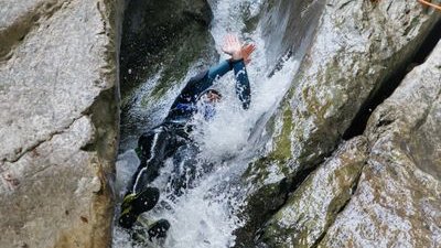 Person sliding down a rocky canyon in water, wearing a wetsuit and helmet; two guides observing.