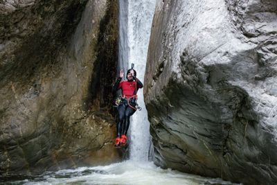 Person in Neoprenanzug und Helm beim Canyoning an einem Wasserfall zwischen felsigen Klippen, mit einem Gurtzeug.