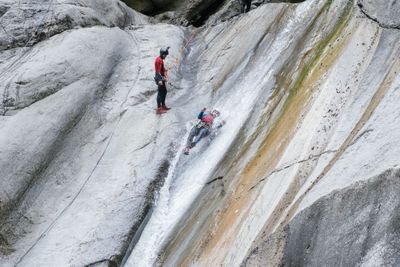 Person rutscht eine nasse Felsfläche hinunter, trägt einen Helm und Klettergurt und wird von einer anderen Person geführt.