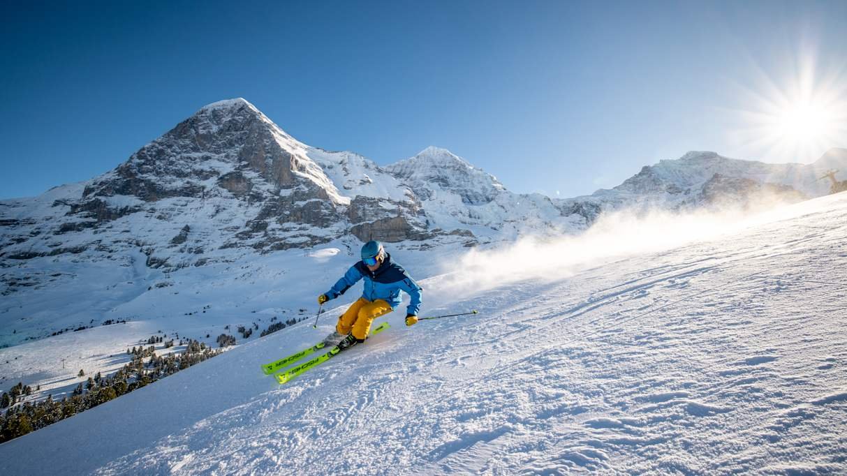 Skifahrer in blauer Jacke und gelber Hose fährt eine verschneite Piste in den Bergen hinunter.