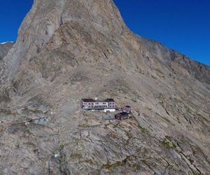 Mountain hut on a rocky slope with a glacier in the background under a clear blue sky.