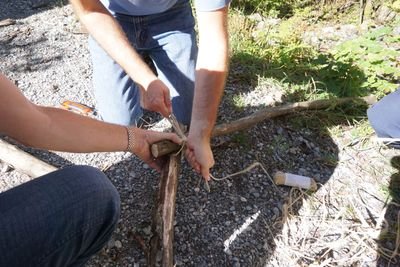 Two people tie rope around wooden sticks on a gravel path outdoors.