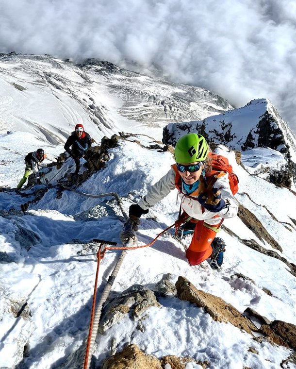Climbers with helmets and harnesses ascend a snowy mountain ridge, using ropes for support.