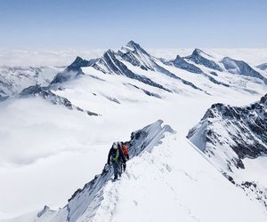 Two climbers in helmets and harnesses traverse a snowy mountain ridge with peaks in the background.
