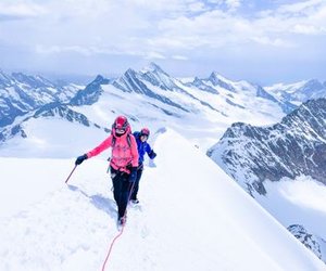Two climbers in helmets and harnesses ascend a snowy mountain ridge with a rope in the Swiss Alps.