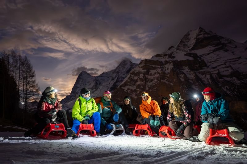 People sitting on sleds in snow at night, wearing winter clothing and headlamps, with mountains behind.