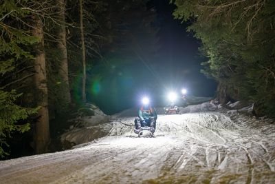 Menschen rodeln nachts auf einem verschneiten Waldweg und tragen Helme mit Stirnlampen.