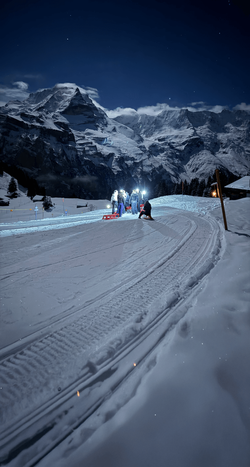 People sledding at night on a snowy mountain trail, wearing winter gear and helmets, with headlamps.