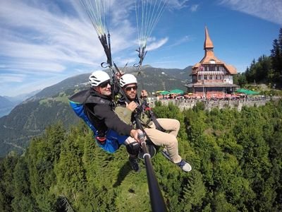 Zwei Personen beim Gleitschirmfliegen über bewaldeten Hügeln, mit Helmen und Gurtzeug, nahe einem Gebäude.