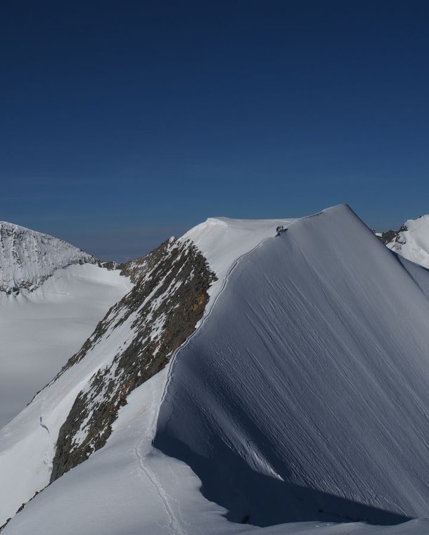 Climbers on a snowy mountain ridge with visible peaks in the background under a clear blue sky.