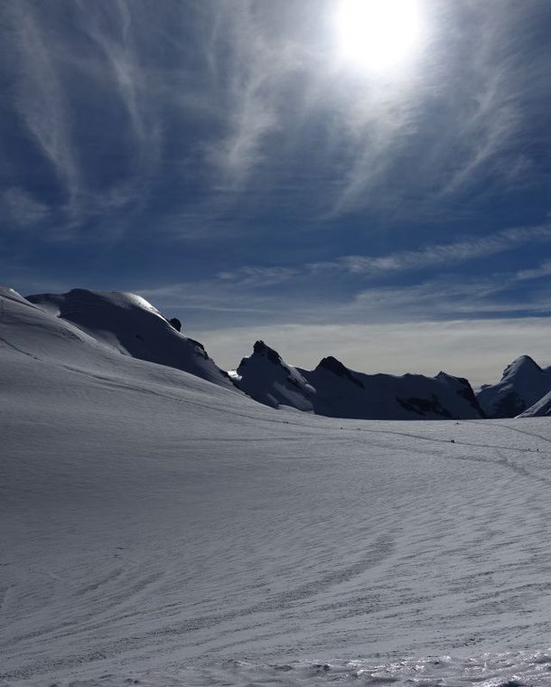 Snow-covered mountain landscape under a bright sun, with distant peaks and ski tracks visible.