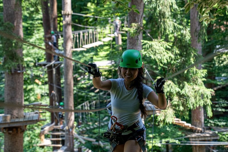 Person trägt einen grünen Helm und ein Geschirr und navigiert einen Seilparcours in einem dichten Waldabenteuerpark.