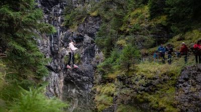 Person auf Seilbrücke in Bergschlucht, gesichert mit Klettergurt; Zuschauer am Rand.