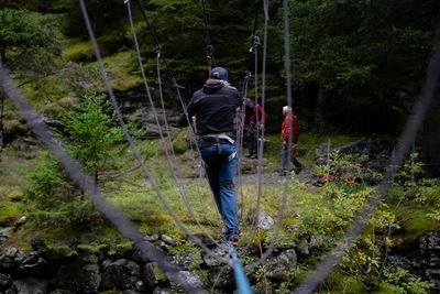 Person auf Seilbrücke im Wald mit Klettergurt und Helm; zwei Personen stehen am Boden.