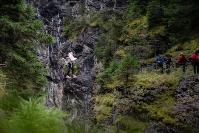 Person auf Seilbrücke in Bergschlucht, gesichert mit Klettergurt; Zuschauer am Rand.