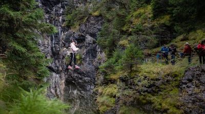 Person auf Seilbrücke in Bergschlucht, gesichert mit Klettergurt; Zuschauer am Rand.