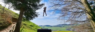 Person with helmet and climbing harness on a rope while ziplining in a forested, hilly landscape under a blue sky.