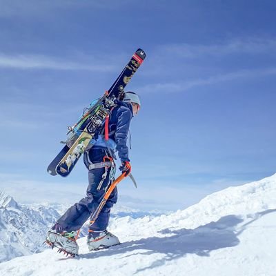 Eine Person geht mit einem Pickel in der Hand, einem Rucksack und aufgebundenen Skis einen schneebedeckten Berg hinauf. Im Hintergrund ist blauer Himmel und eine Bergkette zu erkennen.