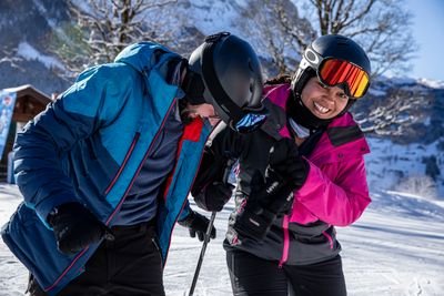 Zwei Personen mit Helmen und Skiausrüstung auf einer schneebedeckten Piste in den Bergen.