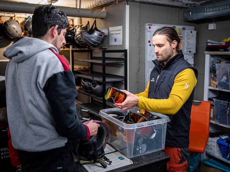 Two people in a gear room, one handing ski goggles to the other, surrounded by helmets.