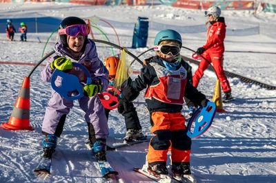 Zwei Kinder mit Helmen und Skiern auf einer Piste, begleitet von einem Skilehrer in roter Kleidung.
