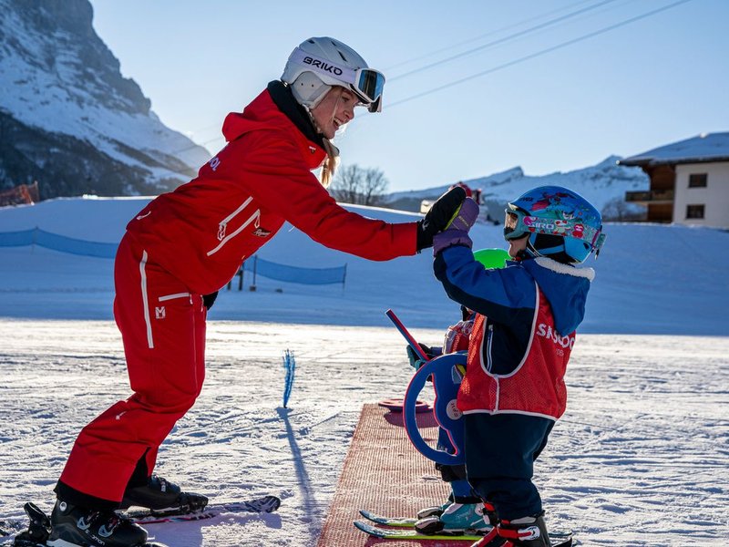 Ski instructor in red gear gives instructions to a child wearing a helmet and skis on a snow-covered slope.