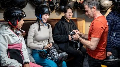 People in helmets sit indoors, preparing for an activity with a guide assisting with gear.