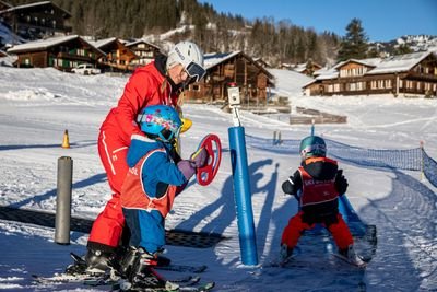 Zwei Kinder mit Helmen und Skiern auf Förderband, begleitet von einem Erwachsenen in roter Skibekleidung.