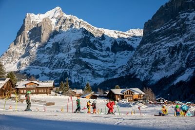 Kinder mit Skiern und Helmen auf Skipiste, begleitet von einem Erwachsenen, vor schneebedeckten Bergen.