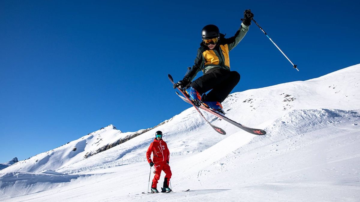 Personen beim Skifahren auf schneebedecktem Berg; eine Person springt, beide mit Helmen und Skiern.