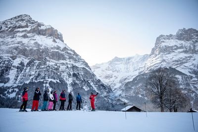 Skifahrer in Helmen und Ausrüstung stehen mit einem Führer auf einem verschneiten Hang, umgeben von schneebedeckten Bergen.