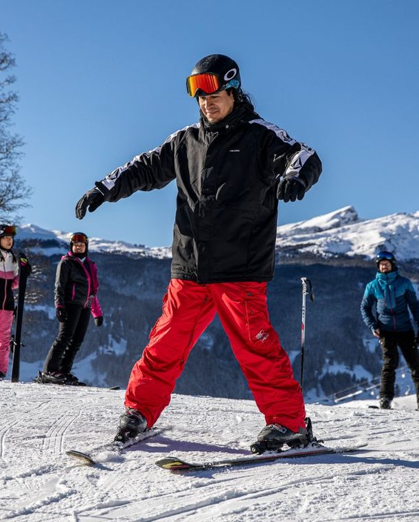 A group of skiers on a snowy mountain slope, wearing helmets and ski gear, with mountains in the background.