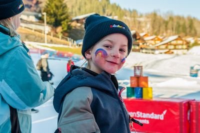 Kind auf Skipiste mit bemaltem Gesicht, Mütze und Winterjacke; verschneite Berge im Hintergrund.